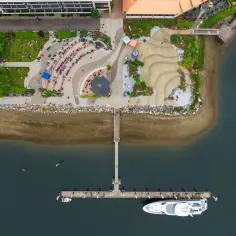 Widok z lotu ptaka na ruchliwy **Olimpia Port** z ludźmi siedzącymi na trawie i łodziami przycumowanymi do nabrzeża.