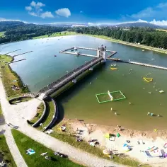 Letnia rekreacja nad jeziorem, z pomostem i wieżą widokową. Woda pełna ludzi, a na brzegu plażowicze cieszą się słońcem. To idealne czarna góra atrakcje dla każdego.