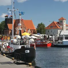 Port na Helu tętni życiem. Widać cumujące łodzie, budynki z czerwonymi dachami i ludzi spacerujących po nabrzeżu.