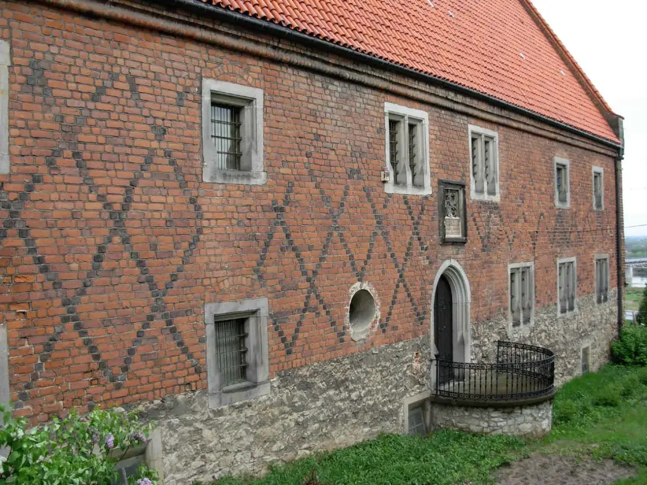 Kamienny, ceglany dom Długosza z charakterystycznym romboidalnym wzorem na ścianie, ozdobiony kamiennymi detalami i balkonikiem.