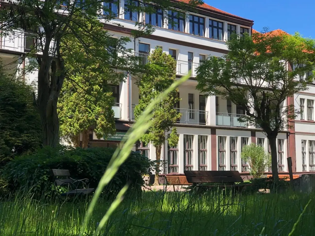 Sanatorium uzdrowiskowe Kołobrzeg-Podczele NFZ otoczone zielenią. Widać ławki w trawie i drzewa.