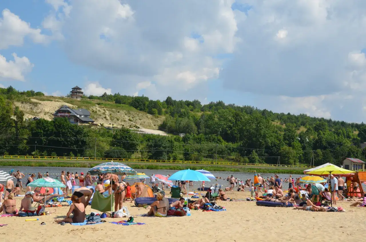 Tłum ludzi odpoczywa na piaszczystej plaży nad zalewem w Krasnobrodzie. W tle zielone wzgórza i budynki.