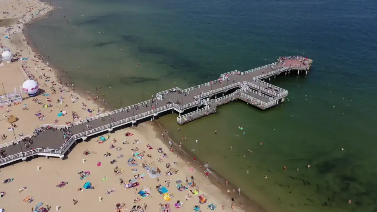 Tłum ludzi wypoczywa na piaszczystej plaży Brzeźno, spacerując po molo lub kąpiąc się w morzu.