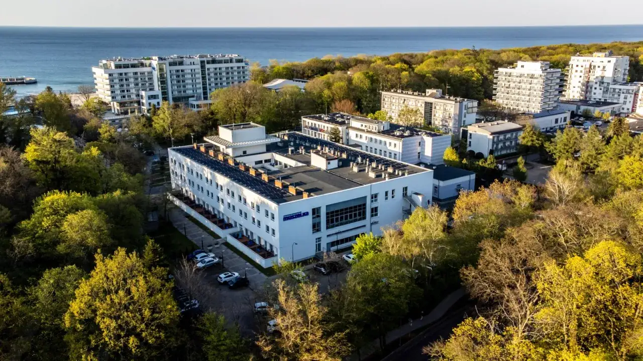 Kołobrzeg sanatorium Muszelka, nowoczesny budynek z panelami słonecznymi, otoczony zielenią, z widokiem na morze.