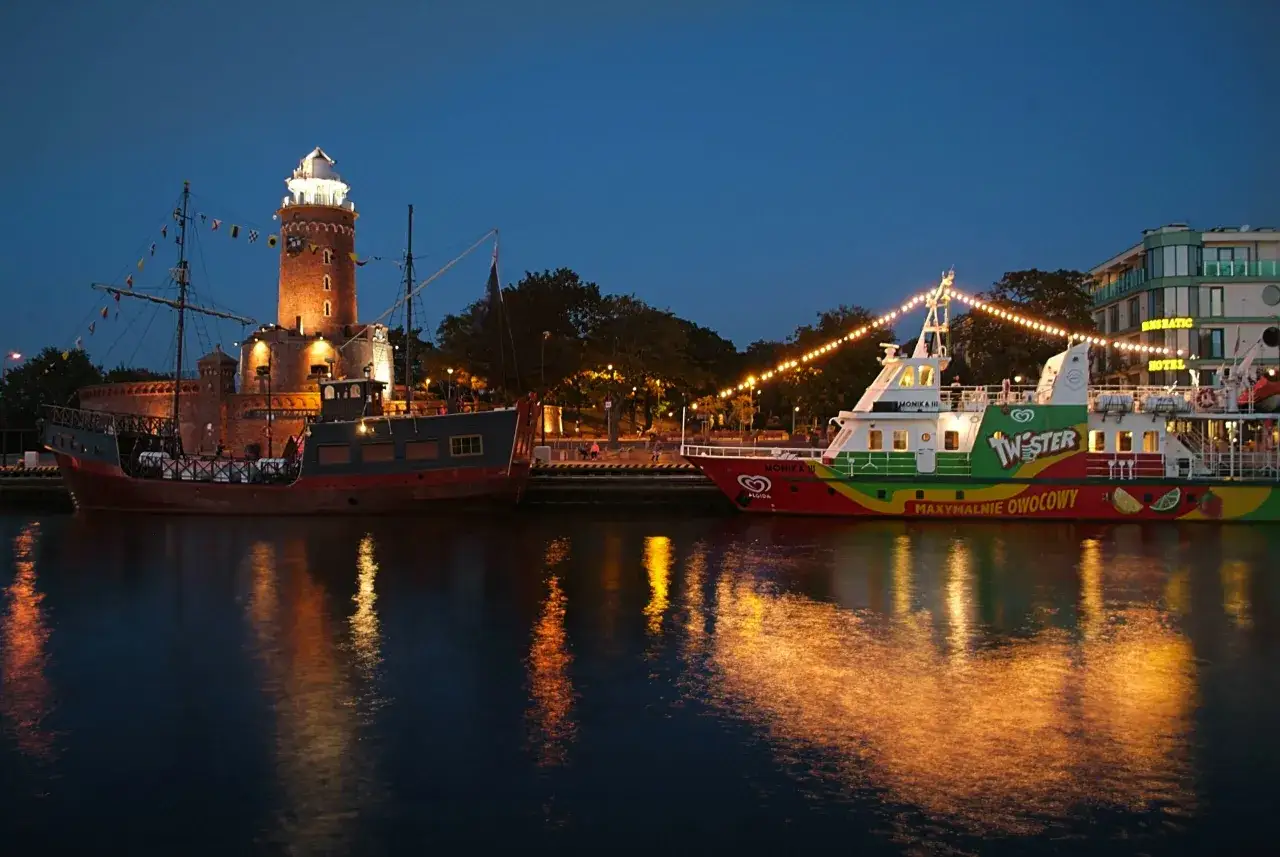 Wieczorny port w Kołobrzegu: zabytkowy żaglowiec i kolorowy statek wycieczkowy przy oświetlonej latarni morskiej.