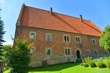 Zabytkowy dom Długosza z cegły, z charakterystycznym dachem i kamiennym cokołem, otoczony zielenią.
