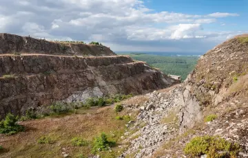 Widok na tarasową kopalnię kwarcu Stanisław. Skarpy skalne z widocznymi warstwami skał, porośnięte trawą i nielicznymi drzewami. W oddali las i błękitne niebo z chmurami.