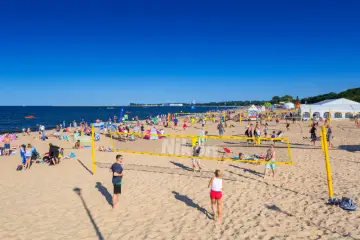 Słoneczna plaża Brzeźno tętni życiem. Ludzie grają w siatkówkę plażową, opalają się i spacerują.
