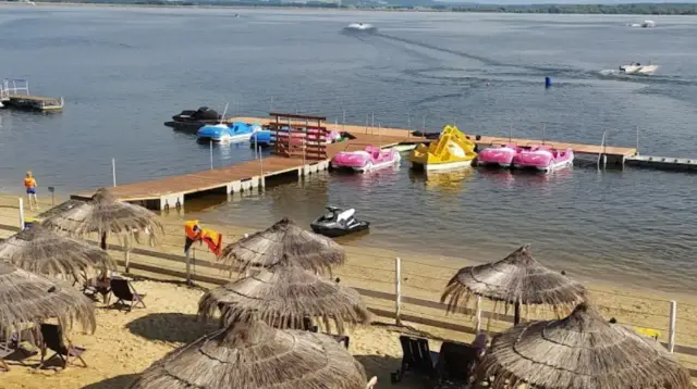 Słoneczna plaża Skorochów z parasolami, leżakami i kolorowymi łódkami wodnymi przy drewnianym pomoście.