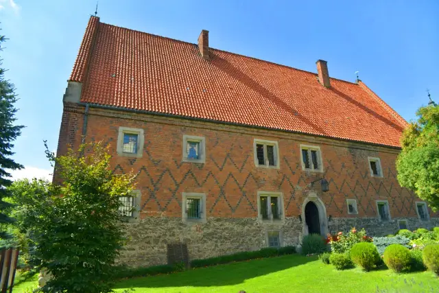 Zabytkowy dom Długosza z cegły, z charakterystycznym dachem i kamiennym cokołem, otoczony zielenią.
